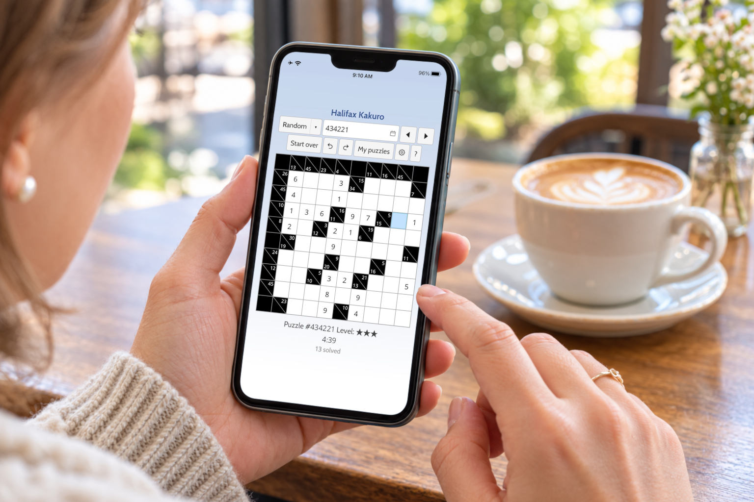 Someone in a caf&eacute; holding a phone with the Halifax Kakuro app open, a cup of coffee beside it